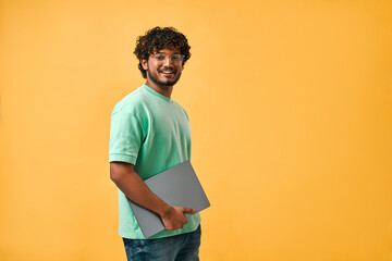 Portrait of handsome curly Indian man in turquoise t-shirt and glasses laughing looking at camera and holding laptop.