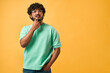 Portrait of a handsome curly-haired Indian man in a turquoise t-shirt and glasses thoughtfully scratching his chin and looking up. The emotion of thoughtfulness, doubt. Copy space.
