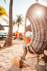 Sticker - Female relaxing on a wicker seat next to a Colorful Michelada Cocktail drink on sandy beach