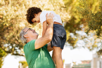 Wall Mural - Glad senior man with beard raise black little boy, have fun, play active game, relax in park, outdoor