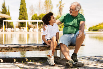 Wall Mural - Cheerful senior man with beard and black little boy sitting on bench, relaxing in park, talk