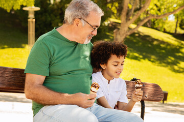 Wall Mural - Cheerful black little boy, senior caucasian man with beard sit on bench, eating ice cream, relax in park