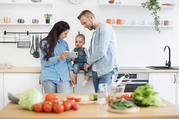 Wall Mural - Happy young family of three in denim outfits looking at mobile screen behind kitchen table at home. Parents and kid searching for online recipe on phone with fresh ingredients for salad in foreground.