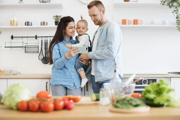 Cheerful caucasian couple and cute baby girl smiling at cell phone screen while leaning against countertop in kitchen. Modern three-person family following top cooking blogger over internet app.