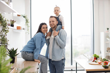 Wall Mural - Portrait of cheerful mom sitting on countertop while snuggling to strong dad with kid on shoulders in dining room. Joyful family of three enjoying pleasant pastime spent in spacious kitchen interior.