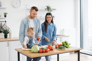 Wall Mural - Handsome father in denim shirt giving tomato to little daughter while pretty mother mastering knife skills at kitchen table. Little infant learning colors and forms while relishing family activities.