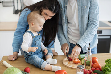 Wall Mural - Cropped view of man using sharp knife with cucumber while small kid watching cautiously at dad's activity. Mindful mother teaching daughter rules of utilizing kitchen tools during food preparation.