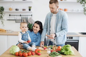 Wall Mural - Portrait of cute little girl sitting on kitchen table while loving father and mother preparing meals for lunch at home. Sweet baby helping parents creating romantic dinner for wedding anniversary.