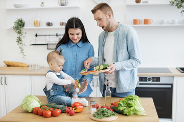 Wall Mural - Shocked father holding cutting board with sliced cucumber while sweet baby girl mixing salad with hands in glass bowl. Curious kid exploring texture and shapes of colorful punch of salad ingredients.