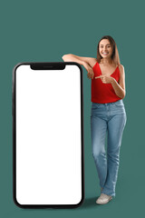 Wall Mural - Happy young woman pointing at big mobile phone with blank screen on green background. Banner for design