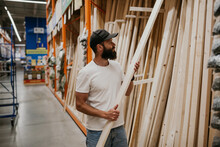 A Young Handsome Man With A Beard In Casual Clothes In A Construction Hypermarket In The Lumber Department Selects Wood Building Materials For The Renovation Of His House.