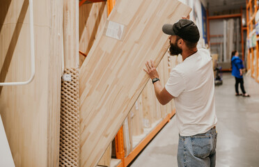 A young handsome man with a beard in casual clothes in a construction hypermarket in the lumber department selects wood building materials for the renovation of his house.