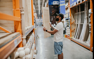 A young handsome man with a beard in casual clothes in a construction hypermarket in the lumber department selects wood building materials for the renovation of his house.