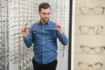 Poster - Satisfied Customer. View of happy young male client wearing new glasses, standing near rack and showcase with eyewear. Smiling man trying on spectacles