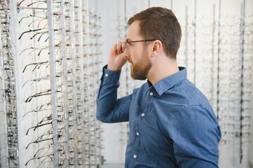 Wall Mural - In Optics Shop. Portrait of male client holding and wearing different spectacles, choosing and trying on new glasses at optical store. Man picking frame for vision correction, closeup.