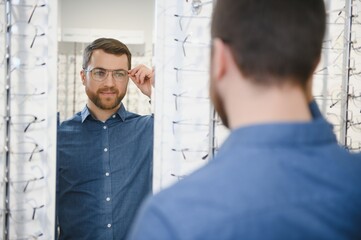 Wall Mural - In Optics Shop. Portrait of male client holding and wearing different spectacles, choosing and trying on new glasses at optical store. Man picking frame for vision correction, closeup.