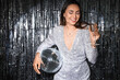 © Pixel-Shot - Smiling fashionable woman in sparkling dress with disco ball and glass of champagne against silver tinsel