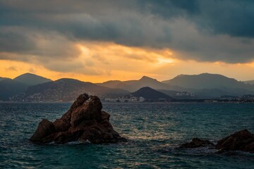 Wall Mural - Rock formation in the sea surrounded by mountains at sunset.