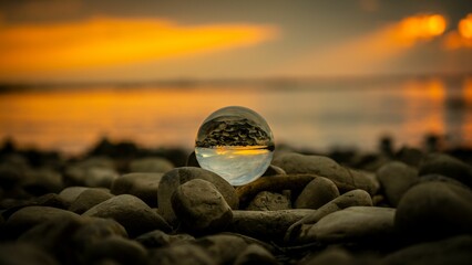 Canvas Print - Low-angle shot of a glass ball on beach rocks reflecting the sea and the sunset light