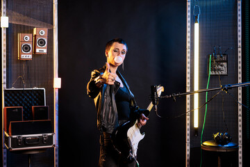 Woman guitarist showing approval sign at camera while performing metal music in sound studio using electric guitar. Rebel performer doing thumbs up gesture, playing rock song at solo performance