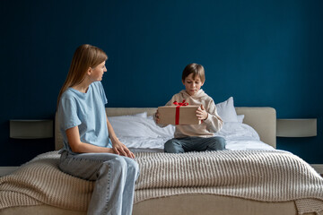 Wall Mural - Happy little kid opening birthday present from mom, excited smiling boy sitting on bed with mother unwrapping wrapped gift box. Loving parent congratulating child at home