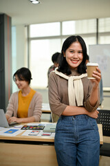 Wall Mural - An attractive Asian female graphic designer stands in the office with a coffee cup in her hand