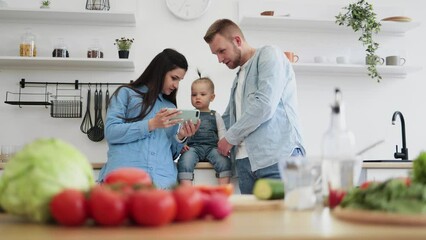 Wall Mural - Happy young family of three in denim outfits looking at mobile screen behind kitchen table at home. Parents and kid searching for online recipe on phone with fresh ingredients for salad in foreground.