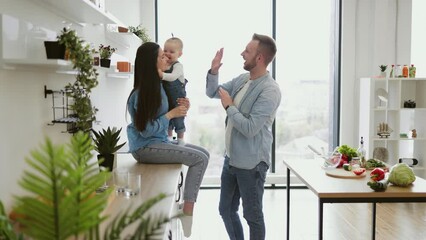 Wall Mural - Beautiful brunette woman sitting on countertop with baby on laps while handsome man standing in middle of room. Funny father playing clapping game with daughter and mother in kitchen interior.