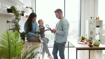 Wall Mural - Beautiful brunette woman sitting on countertop with baby on laps while handsome man standing in middle of room. Funny father playing using kitchen utensils with daughter and mother in kitchen interior