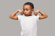 Studio portrait of cute strong muscled little african american kid showing biceps of both arms, looking aside, feeling proud with his strength and endurance, dressed in white mockup t-shirt