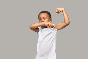 Wall Mural - Handsome sporty athletic boy of 7 years old standing against gray studio background in white mockup t-shirt showing muscled bicep as result of daily workouts. Healthy childhood, fitness kids