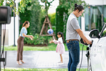 Focus image of progressive man charging electric car from home charging station with blur mother and daughter playing together in the background.