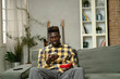 © JustLife - Handsome African man using smartphone while sitting on a sofa in cozy living room. Young man typing a message.