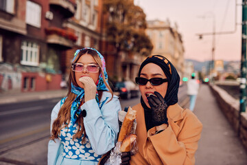 Couple woman one wearing a hijab and a modern yet traditional dress, and the other in a blue dress and scarf, walking together through the city at sunset. One carries a bouquet and bread, while the