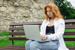 Young red-hair female student using her laptop sitting on the bench in park