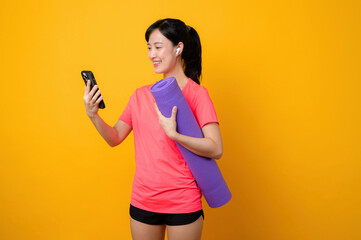 Wall Mural - Portrait young asian sports fitness woman happy smile wearing pink sportswear and smartphone doing exercise training workout against yellow studio background. technology wellness lifestyle concept.