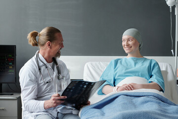 Happy young woman with headscarf sitting in bed and talking to her doctor with brain MRI scan results during medical treatment course