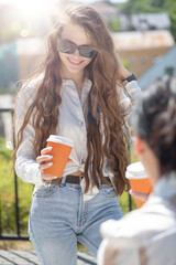 Lifestyle concept.Two beautiful women having fun in city during sunny summer day. Models smiling, talking with each other and drinking coffee. Sun flare effect applied. Image contains motion blur