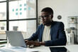 © pressmaster - Young serious African American entrepreneur typing on laptop keyboard while sitting by workplace in office and working over new project