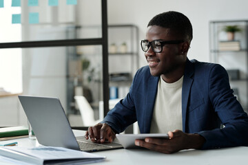 Aufkleber - Young smiling businessman in eyeglasses looking at laptop screen while analyzing online data or communicating in video chat in office
