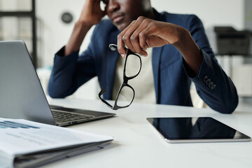 Aufkleber - Focus on hand of young pensive African American businessman holding eyeglasses while sitting by desk with laptop and tablet in office
