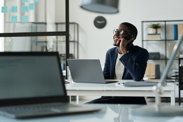 Aufkleber - Happy young African American broker talking to client on smartphone while sitting by workplace in front of laptop in openspace office