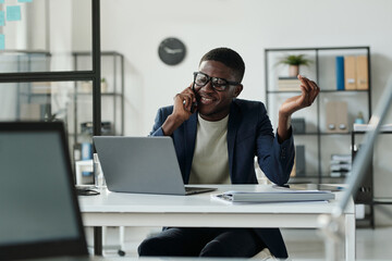Aufkleber - Young cheerful broker talking to colleague on mobile phone while sitting by workplace and looking through financial data on laptop screen