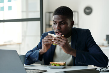 Aufkleber - Young hungry office worker in formalwear and eyeglasses eating sandwich and looking at laptop screen while watching online video by lunch