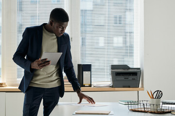 Aufkleber - Young serious businessman with tablet standing by desk with financial documents in openspace office and looking through papers