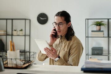 Aufkleber - Young serious chief executive officer in eyeglasses and beige shirt talking on smartphone and looking through online data on tablet screen