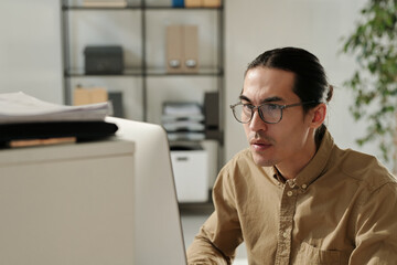 Aufkleber - Young serious analyst in eyeglasses and beige shirt sitting by workplace in front of computer and looking through online data on screen