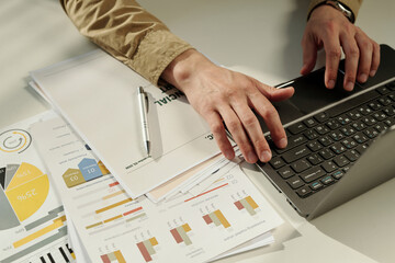 Hands of young male analyst typing on laptop keyboard by workplace while analyzing graphic financial data and entering results in database