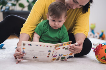 Caring mother showing focused little son book with colorful pictures of living creatures sitting on soft rug in nursery woman educating baby boy at home