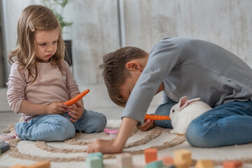 Two kids one toddler girl and one preschooler boy feeding a white rabbit with carrots at home, early child development and unusuall bonding concept